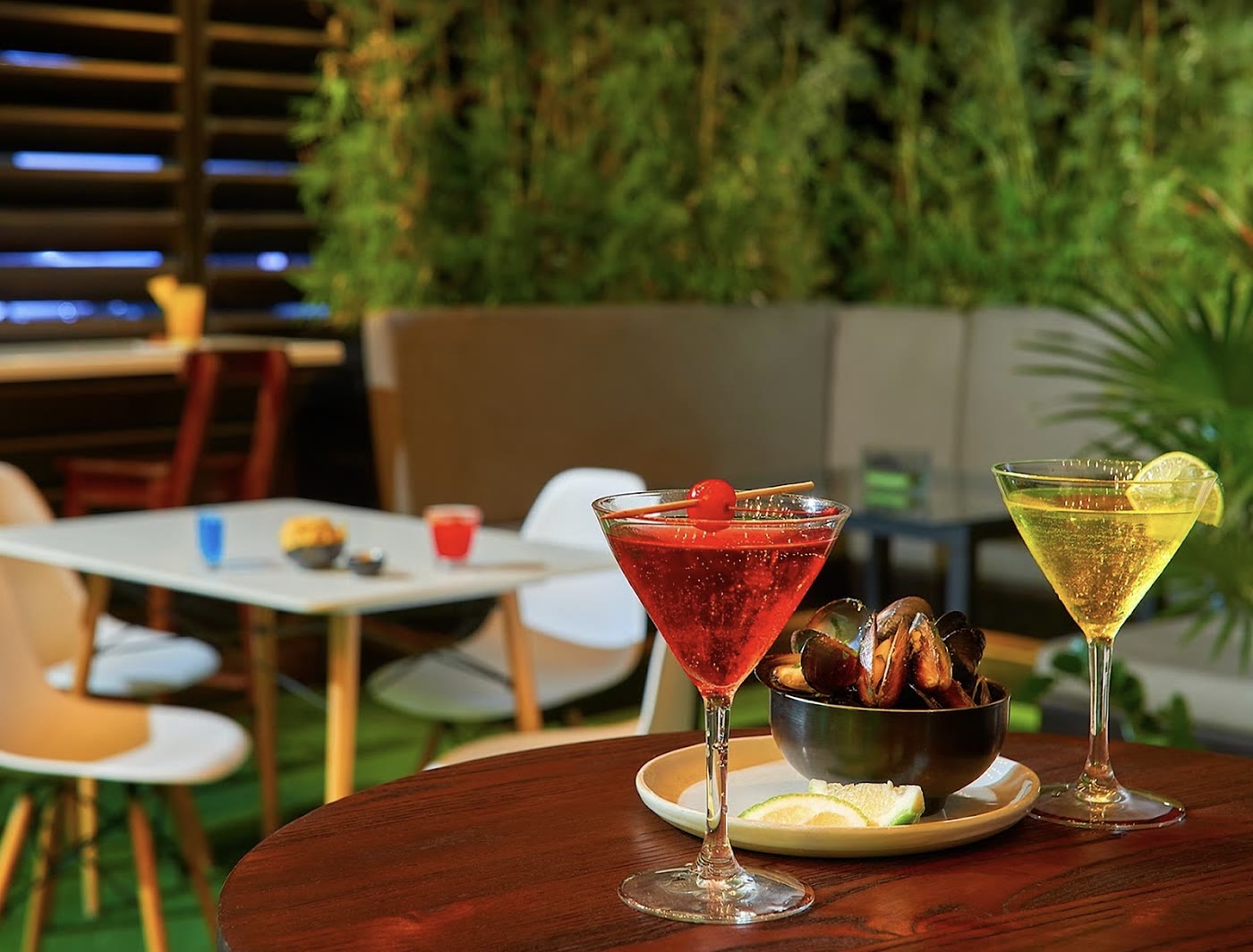 Two martinis, one red and one pale yellow, beside a bowl of mussels on a round wooden table, with a green plant wall behind.