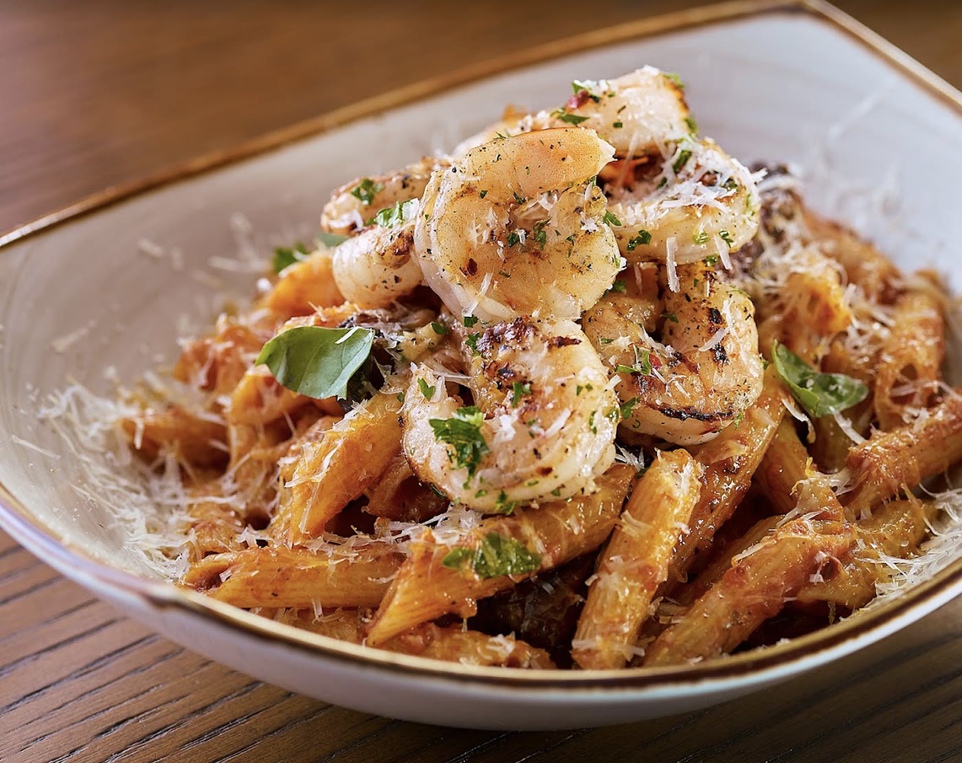 A shallow bowl of penne in tomato sauce, topped with grilled shrimp, shaved parmesan and basil.